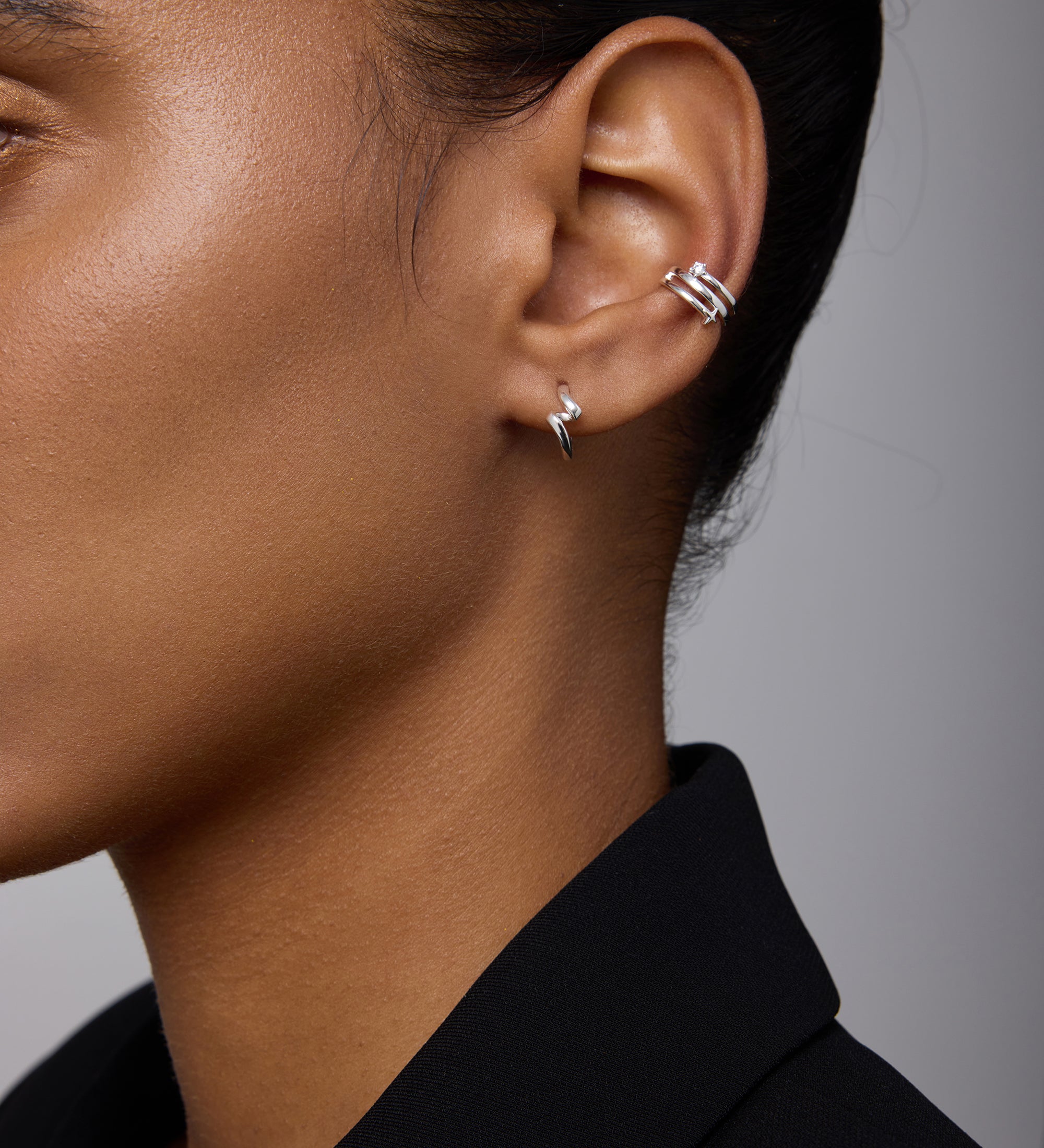Close-up of a person’s ear wearing Ribbon Twist Huggies in Silver on the lobe and another silver huggie on the upper ear. The person has smooth skin and is dressed in a black blazer.