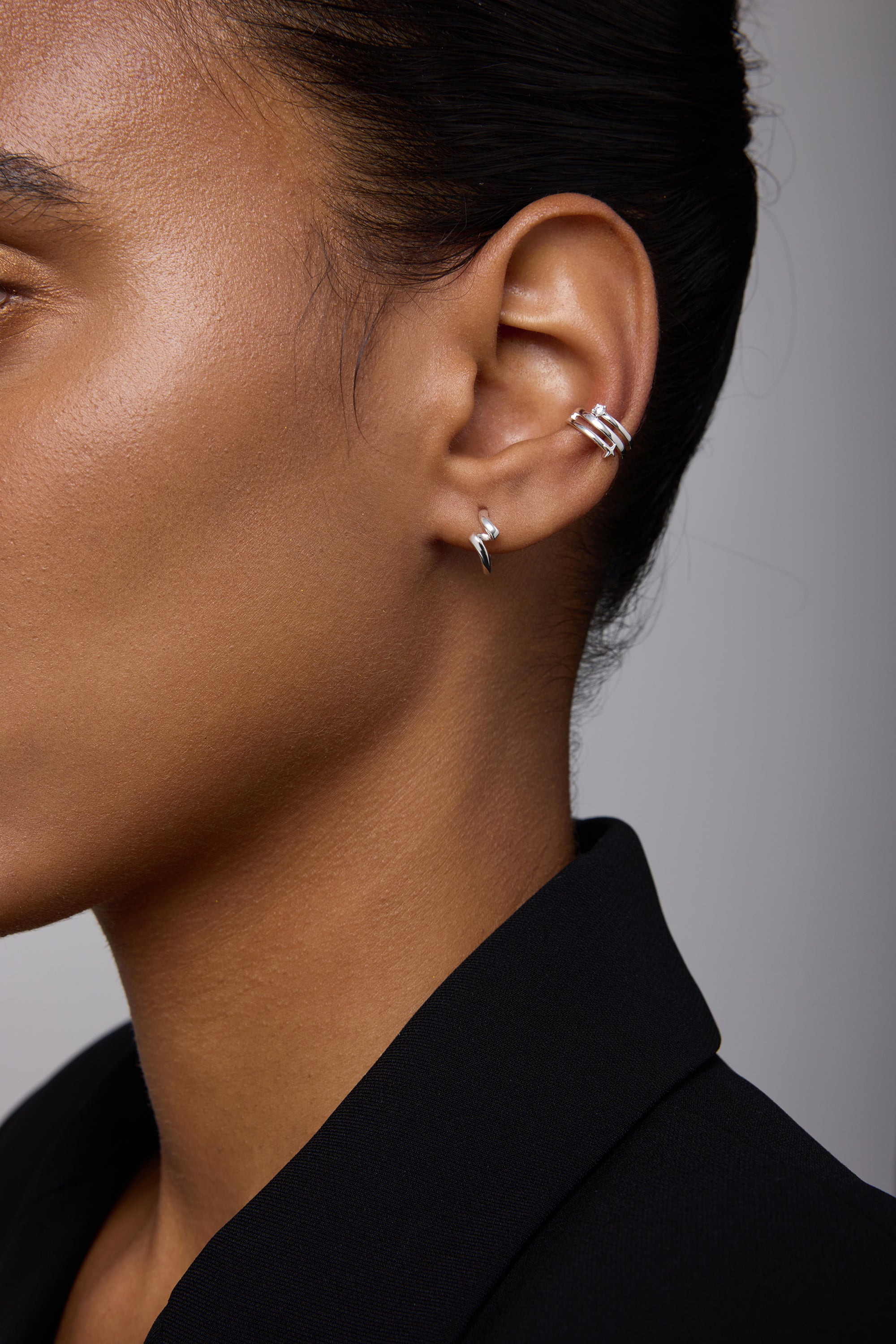 Close-up of a person’s ear wearing Ribbon Twist Huggies in Silver on the lobe and another silver huggie on the upper ear. The person has smooth skin and is dressed in a black blazer.