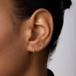 A close-up of an ear with four piercings displays small sustainable gold earrings, turquoise studs, and a 14K Recycled Solid Yellow Gold Rope Huggie Earring. The person has dark hair pulled back and wears a black top.