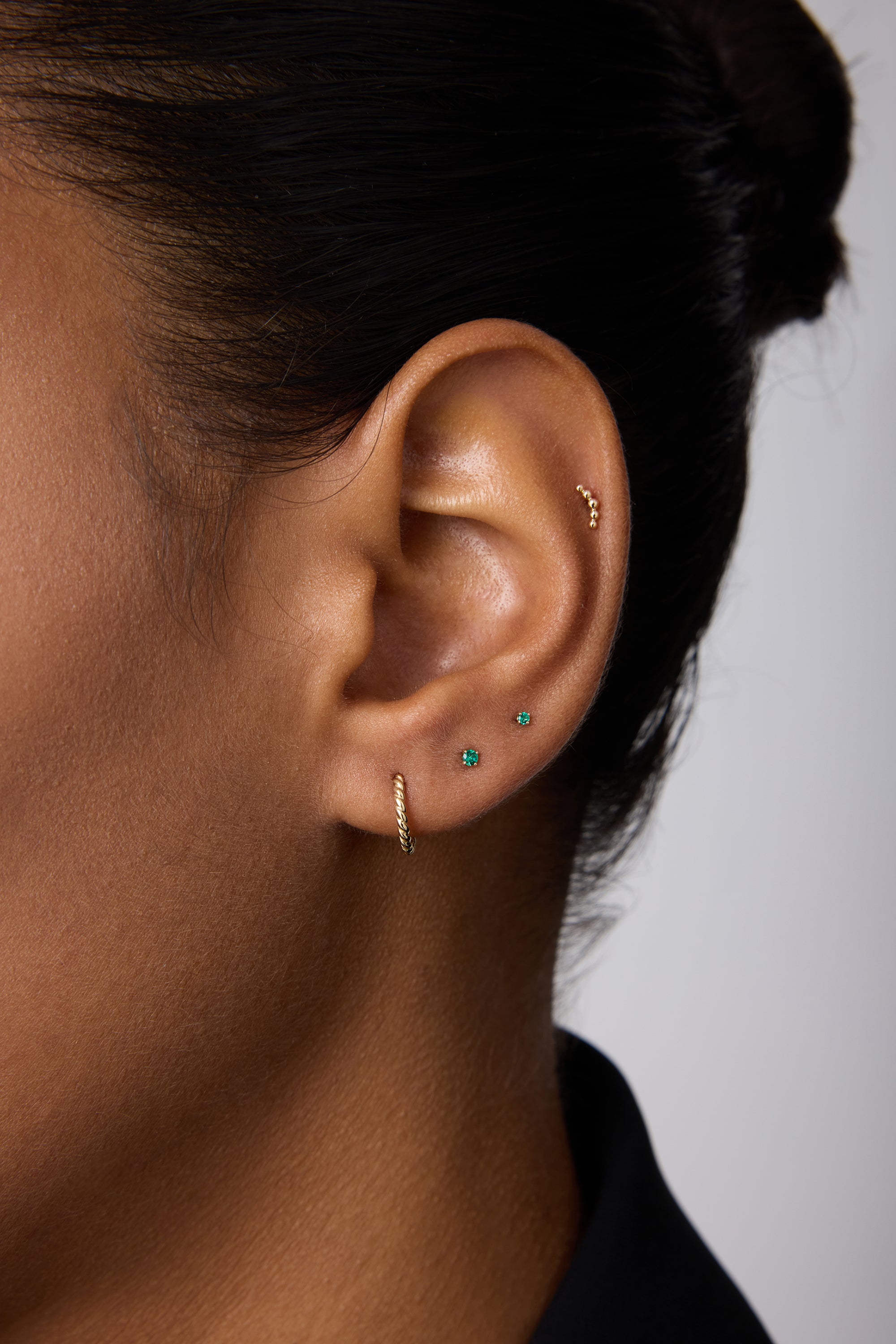 A close-up of an ear with four piercings displays small sustainable gold earrings, turquoise studs, and a 14K Recycled Solid Yellow Gold Rope Huggie Earring. The person has dark hair pulled back and wears a black top.