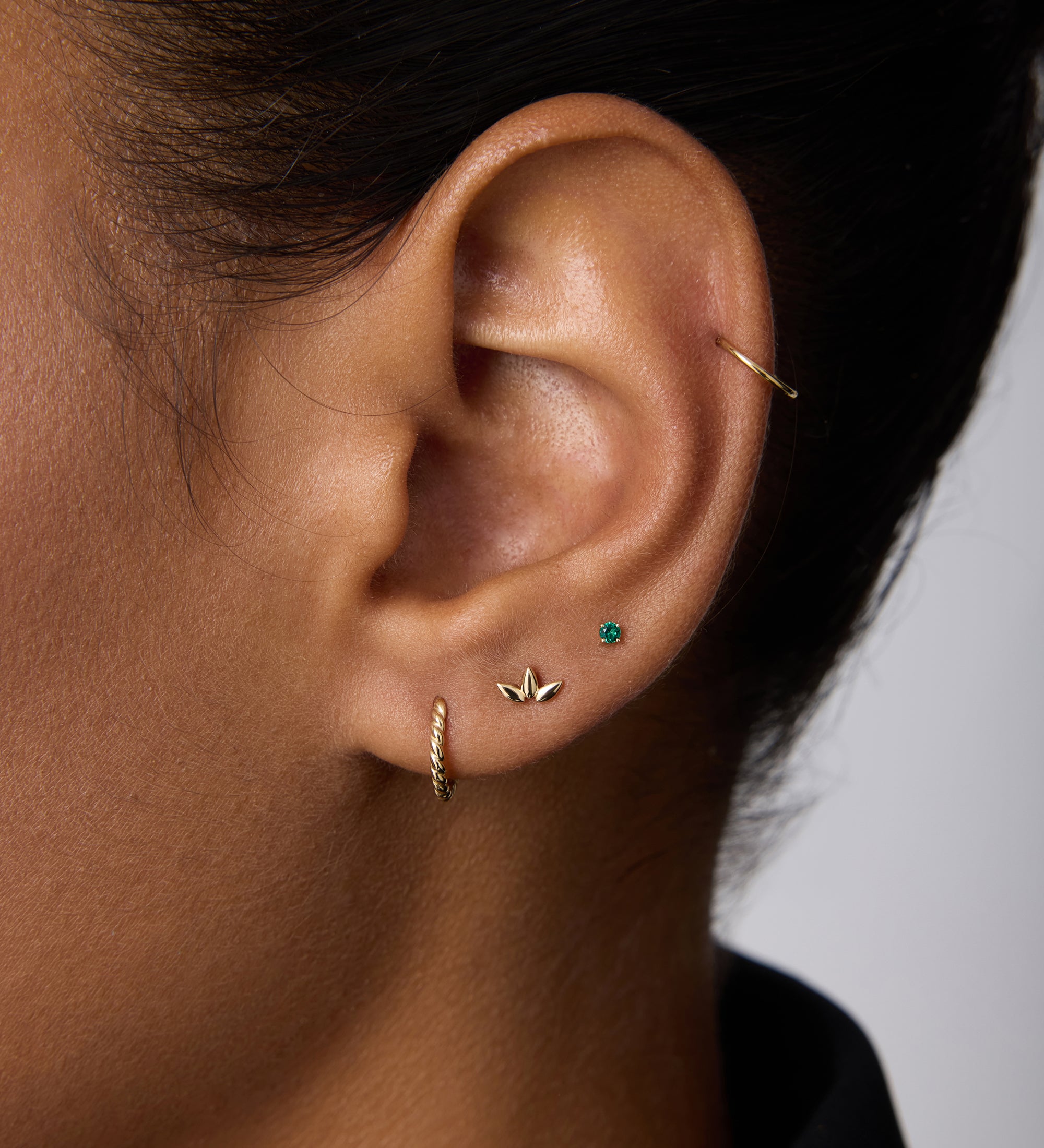 A close-up of a person’s ear with short dark hair wearing a black top, adorned with a 14K Solid Yellow Gold 20G Mini Clicker in the cartilage alongside other gold and turquoise studs.