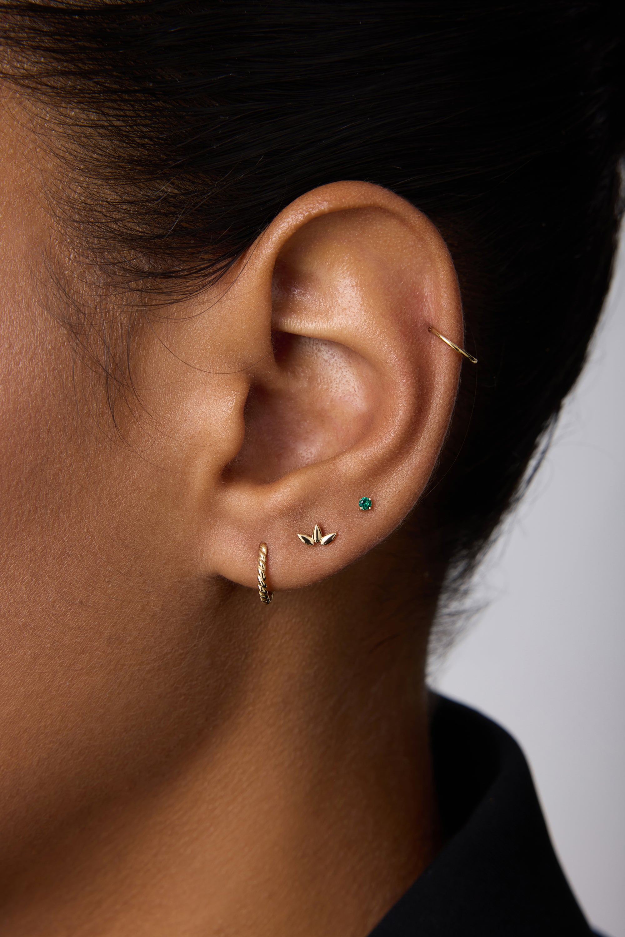 A close-up of a person’s ear with short dark hair wearing a black top, adorned with a 14K Solid Yellow Gold 20G Mini Clicker in the cartilage alongside other gold and turquoise studs.