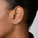 A close-up of a woman with dark skin wearing five silver earrings, including the 14K Solid White Gold Key Flat Back Stud on her upper ear. She also wears a small lobe hoop, three shaped studs above it, and a black collared shirt.