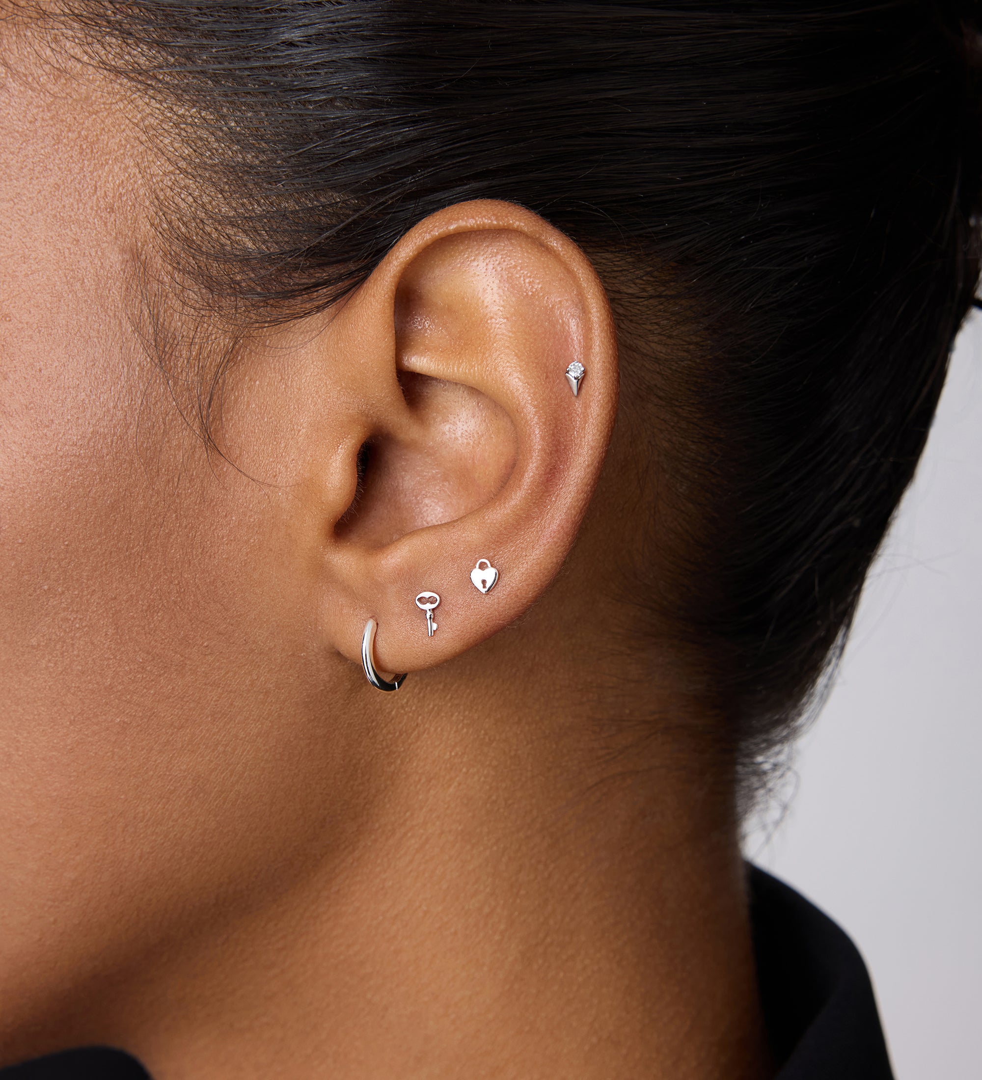A close-up of a woman with dark skin wearing five silver earrings, including the 14K Solid White Gold Key Flat Back Stud on her upper ear. She also wears a small lobe hoop, three shaped studs above it, and a black collared shirt.