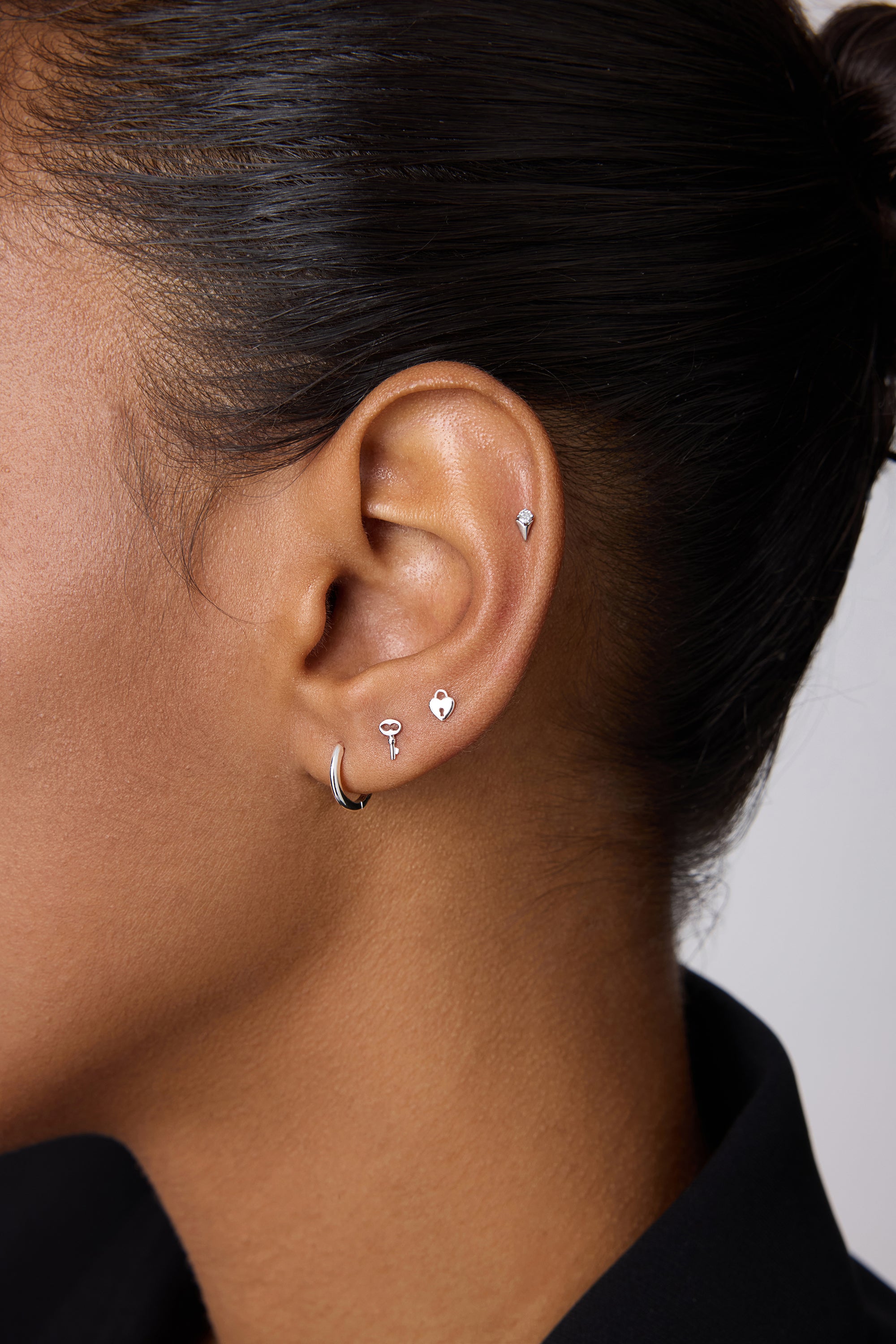 A close-up of a woman with dark skin wearing five silver earrings, including the 14K Solid White Gold Key Flat Back Stud on her upper ear. She also wears a small lobe hoop, three shaped studs above it, and a black collared shirt.