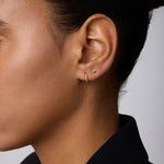 Close-up of a person’s ear adorned with the 14K Solid Yellow Gold Shell Flat Back Stud and a thin gold hoop. The person has smooth skin, dark hair pulled back, and wears a black collared garment.