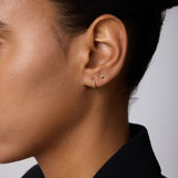 Close-up of a person’s ear adorned with the 14K Solid Yellow Gold Shell Flat Back Stud and a thin gold hoop. The person has smooth skin, dark hair pulled back, and wears a black collared garment.