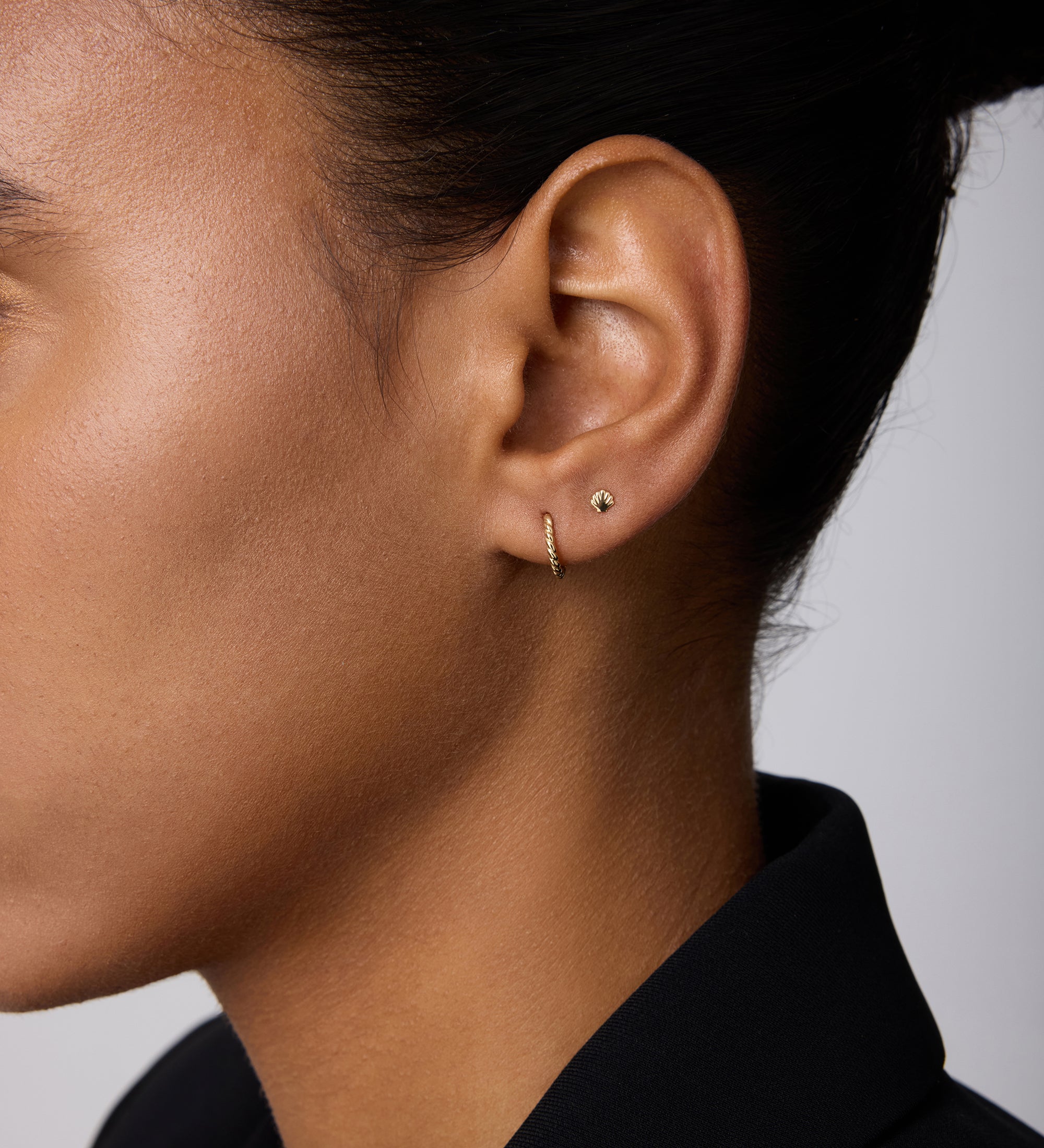 Close-up of a person’s ear adorned with the 14K Solid Yellow Gold Shell Flat Back Stud and a thin gold hoop. The person has smooth skin, dark hair pulled back, and wears a black collared garment.
