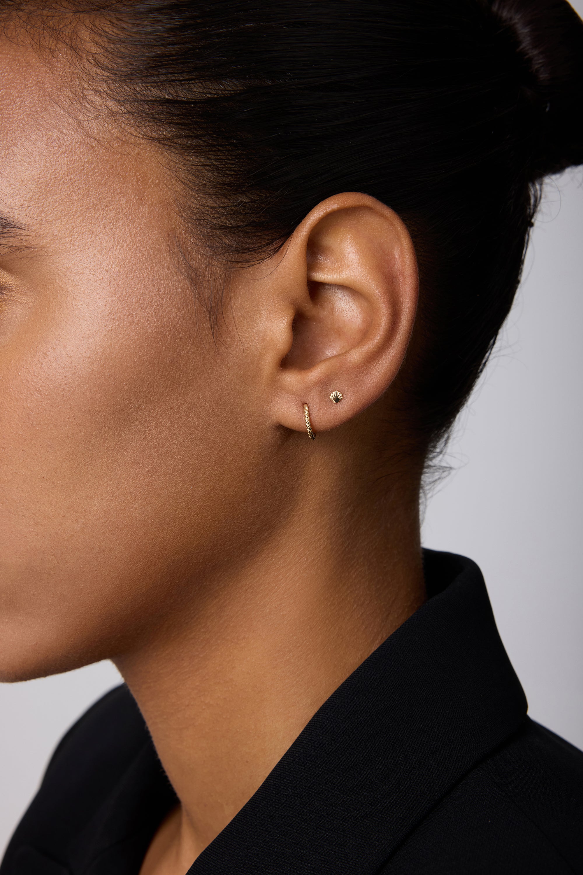 Close-up of a person’s ear adorned with the 14K Solid Yellow Gold Shell Flat Back Stud and a thin gold hoop. The person has smooth skin, dark hair pulled back, and wears a black collared garment.