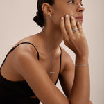A woman with her hair in a low bun wears a black sleeveless top and elegant S925 sterling silver jewelry, including the Bamboo Chain Bracelet in Silver. She rests her chin on her hand and gazes thoughtfully to the side.