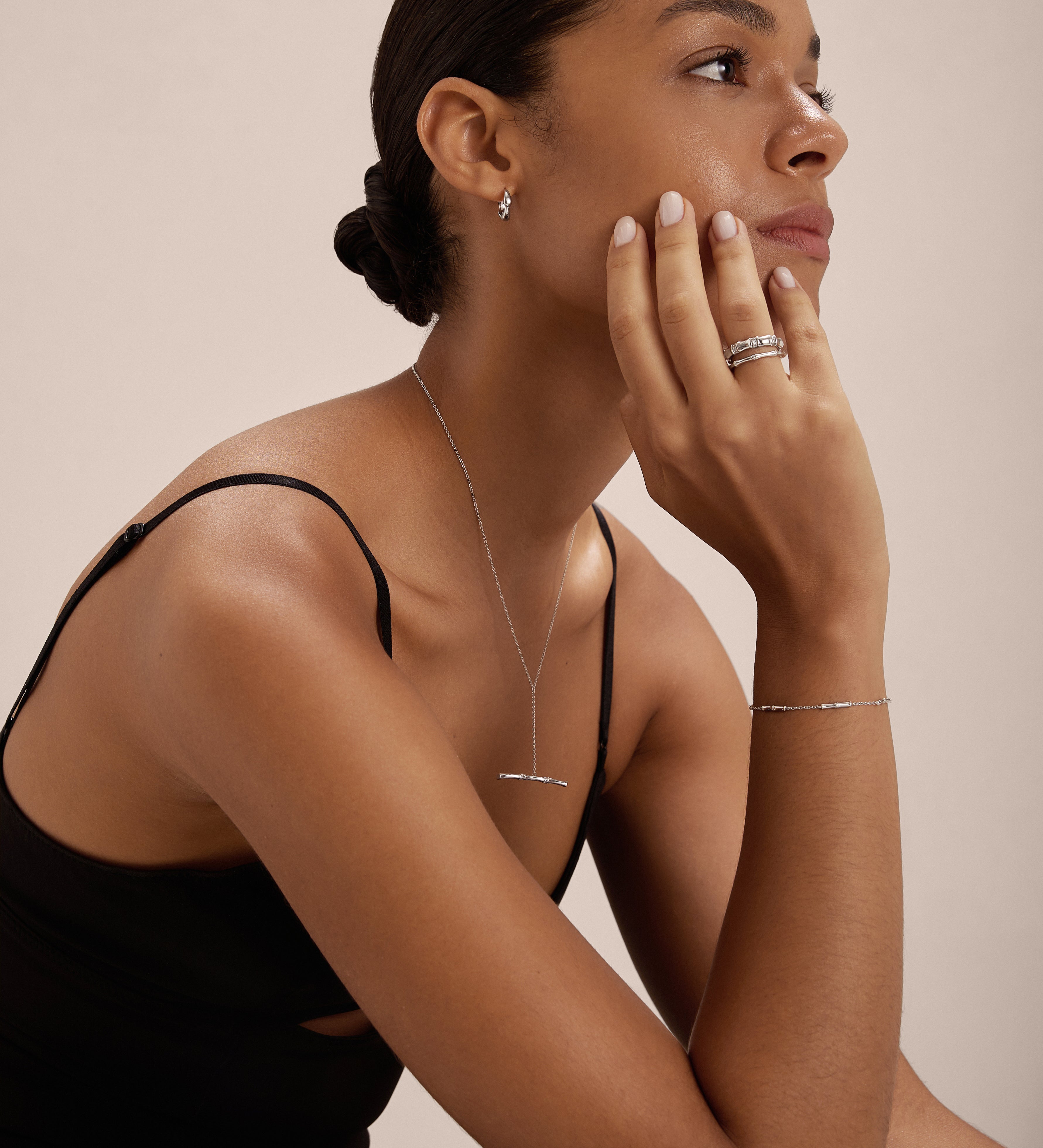 A woman with her hair in a low bun wears a black sleeveless top and elegant S925 sterling silver jewelry, including the Bamboo Chain Bracelet in Silver. She rests her chin on her hand and gazes thoughtfully to the side.