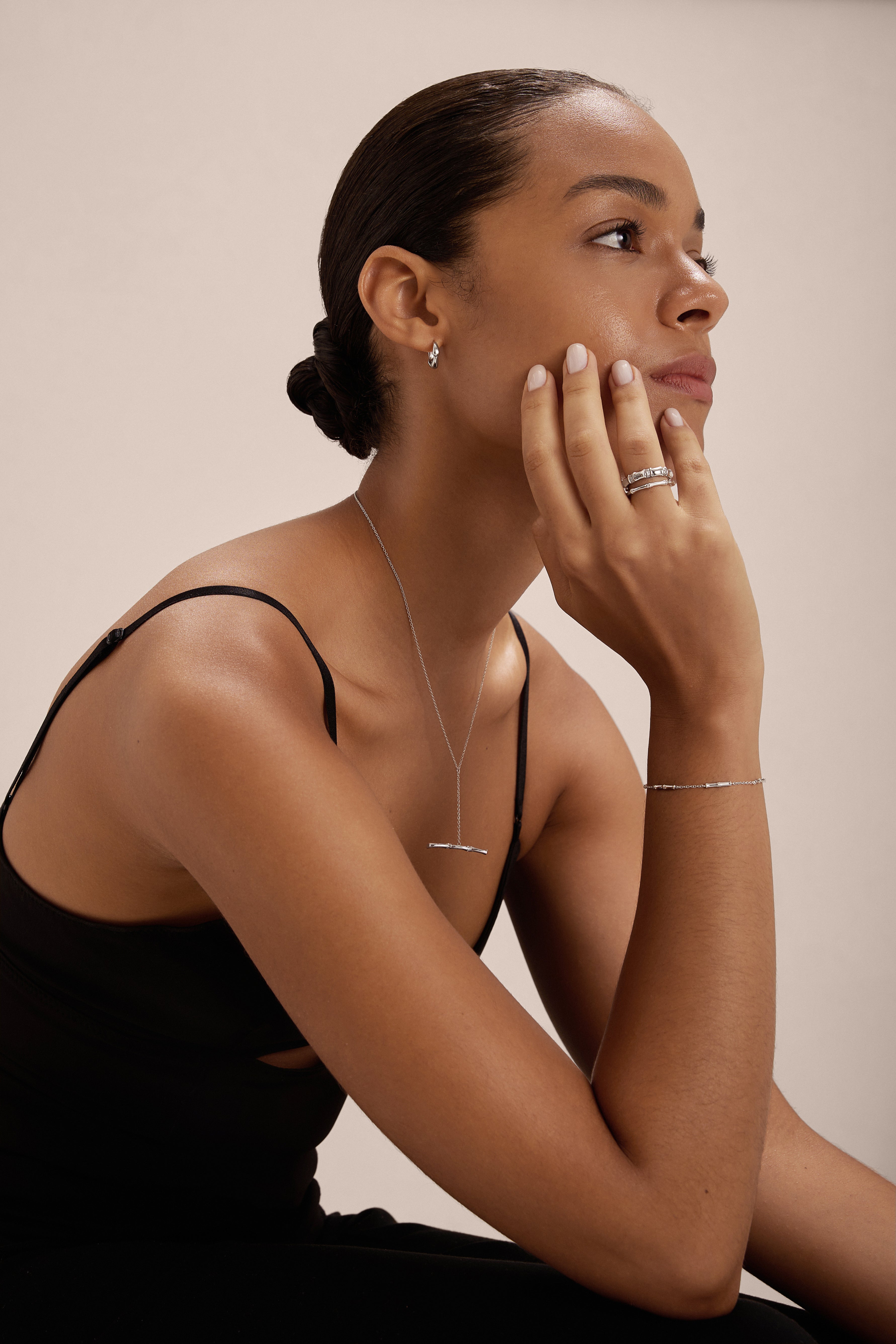 A woman with her hair in a low bun wears a black sleeveless top and elegant S925 sterling silver jewelry, including the Bamboo Chain Bracelet in Silver. She rests her chin on her hand and gazes thoughtfully to the side.