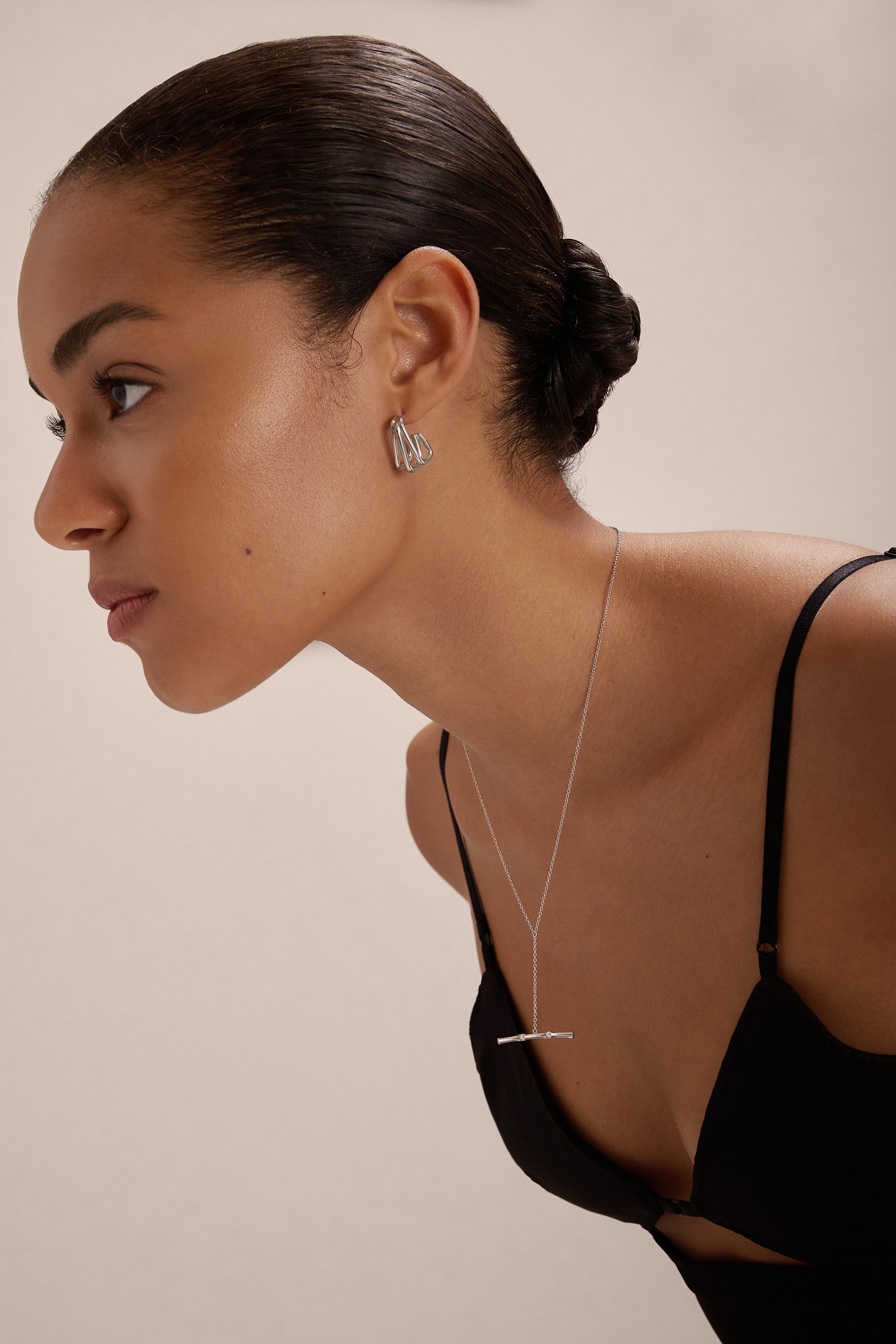 A woman with smooth, pulled-back hair wears a black spaghetti strap top, silver drop earrings, and the Bamboo Chain Y-Bar Lariat Necklace in Silver. She leans forward and looks to the side against a neutral background.