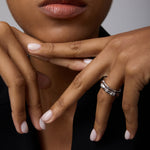 A close-up of a woman with natural lips in black attire, resting her chin on posed hands with pale pink nails, wearing two Bamboo Chunky Crystal Rings in Silver—crafted from recycled sterling silver with rectangular details.