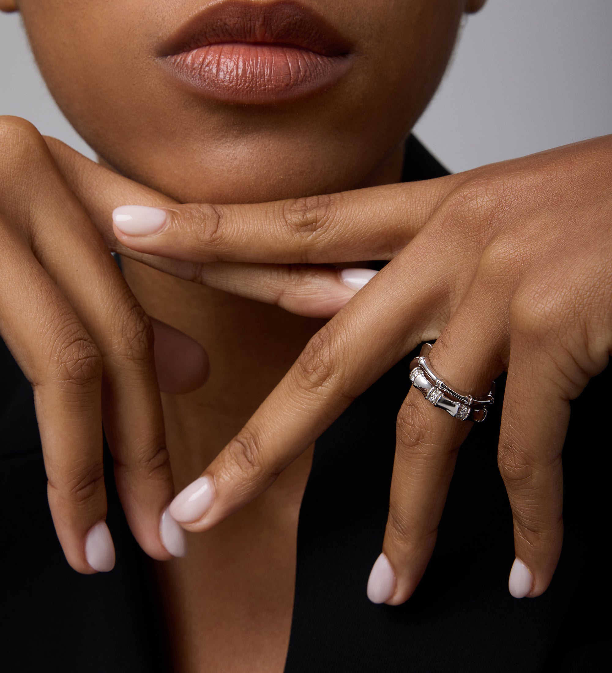 A close-up of a woman with natural lips in black attire, resting her chin on posed hands with pale pink nails, wearing two Bamboo Chunky Crystal Rings in Silver—crafted from recycled sterling silver with rectangular details.