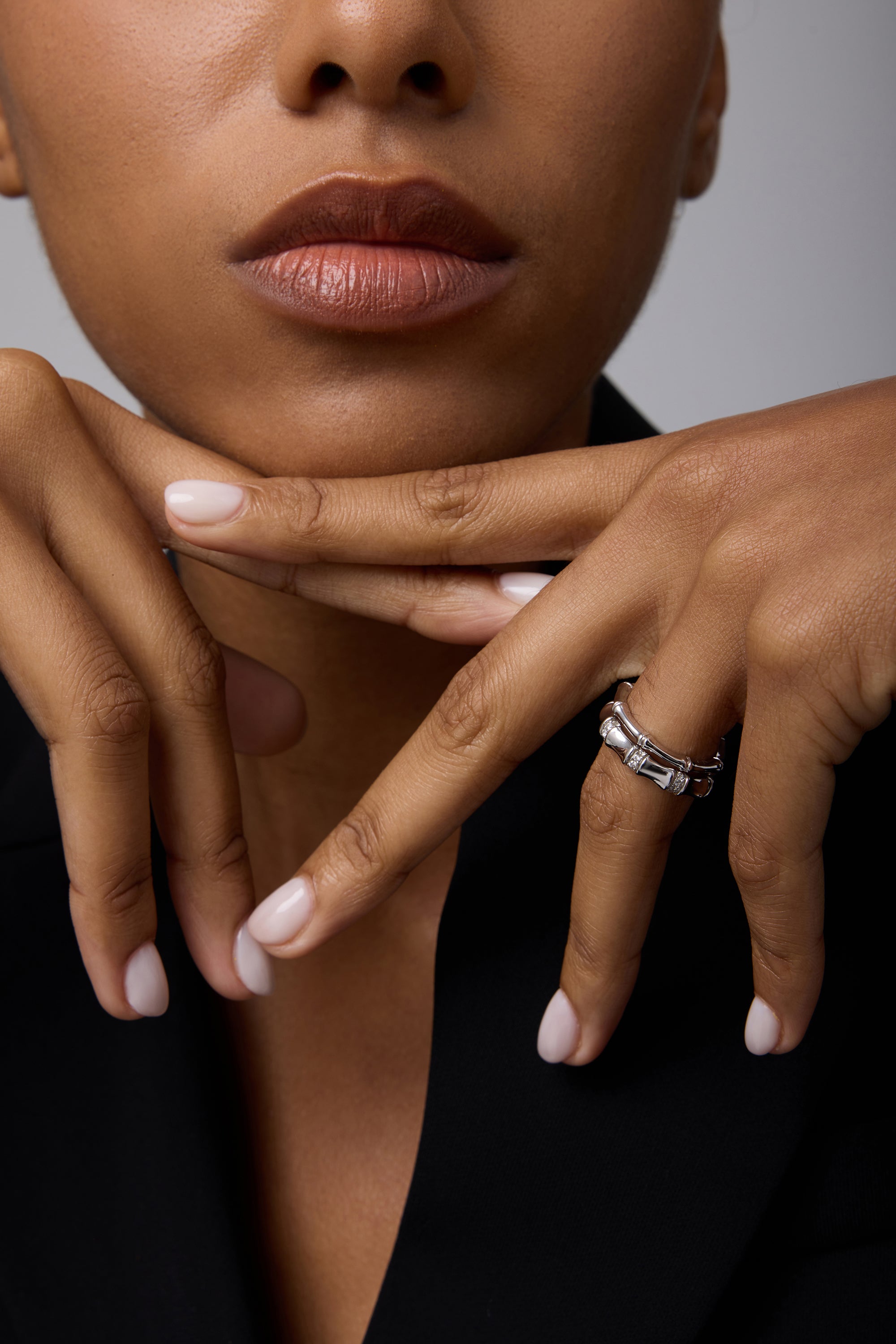 A close-up of a woman with natural lips in black attire, resting her chin on posed hands with pale pink nails, wearing two Bamboo Chunky Crystal Rings in Silver—crafted from recycled sterling silver with rectangular details.