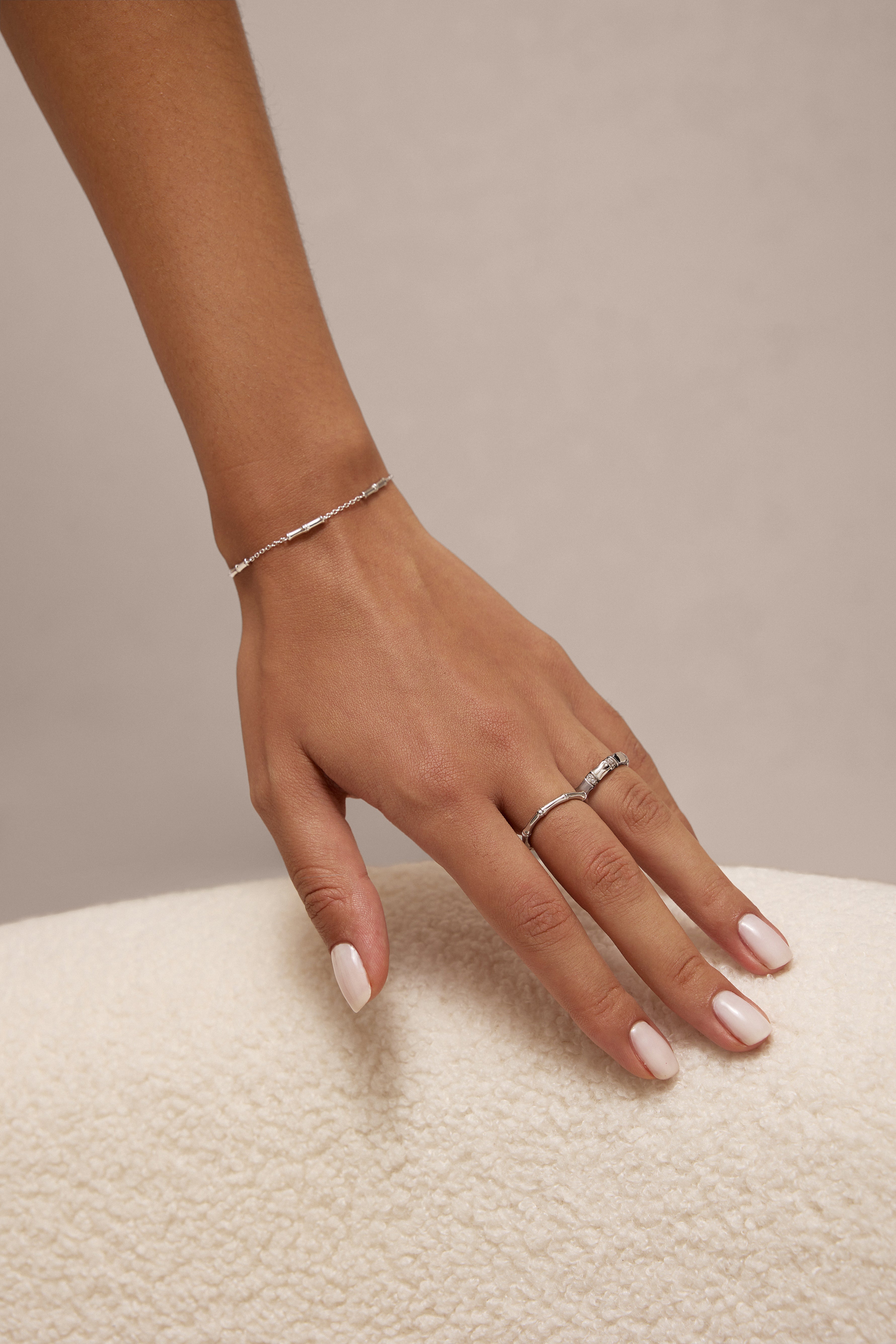 A close-up of a womans light pink manicured hand wearing the Bamboo Ring in Silver—a minimalist piece crafted from recycled sterling silver—plus another simple ring and a delicate silver bracelet, resting on a textured white surface.