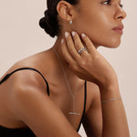 A woman with smooth skin and dark hair in a low bun wears a black sleeveless top and silver jewelry, including earrings, rings, a bracelet, and the Bamboo Chain Y-Bar Lariat Necklace in Silver. She poses in profile with her hand on her chin.