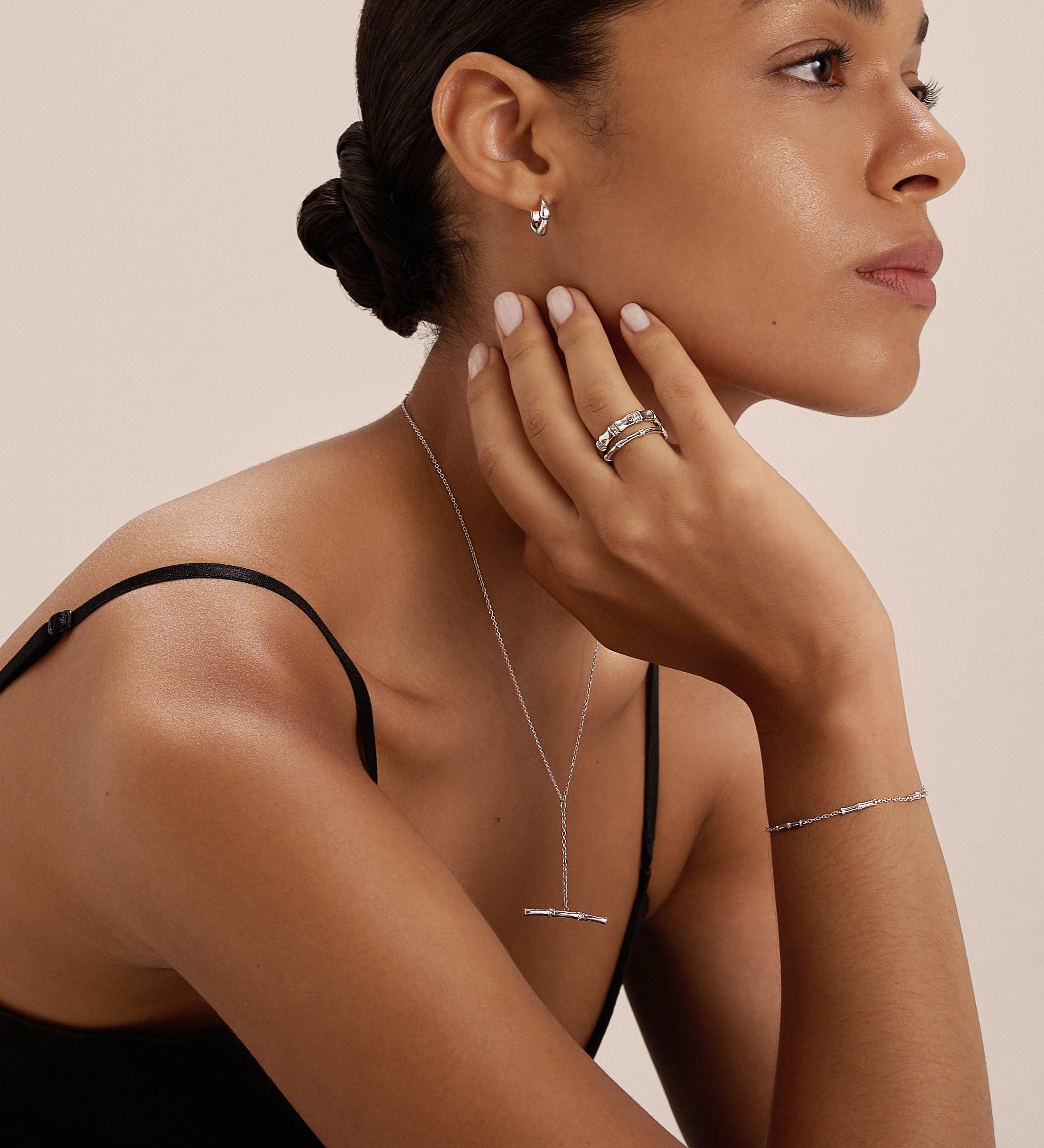 A woman with smooth skin and dark hair in a low bun wears a black sleeveless top and silver jewelry, including earrings, rings, a bracelet, and the Bamboo Chain Y-Bar Lariat Necklace in Silver. She poses in profile with her hand on her chin.