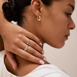 A woman with smooth skin and dark hair in a low bun wears gold hoop earrings and a Charlotte Eternity Ridged Ring in Silver, her hand resting on her neck. She is dressed in a cream top with red stripes and has light pink manicured nails.