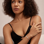 A woman with curly hair in a black strappy top sits with one arm crossed over her chest, wearing the Gisèle Cuban Link Bar Bracelet in Silver and a matching ring. She gazes thoughtfully to the side against a neutral background.
