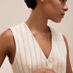 A woman with dark hair in a low bun models the Gisèle Cuban Link Bar Bracelet in Silver, paired with minimalist silver jewelry—hoop earrings, a bar necklace, and a simple ring—while wearing a cream pinstriped sleeveless top.