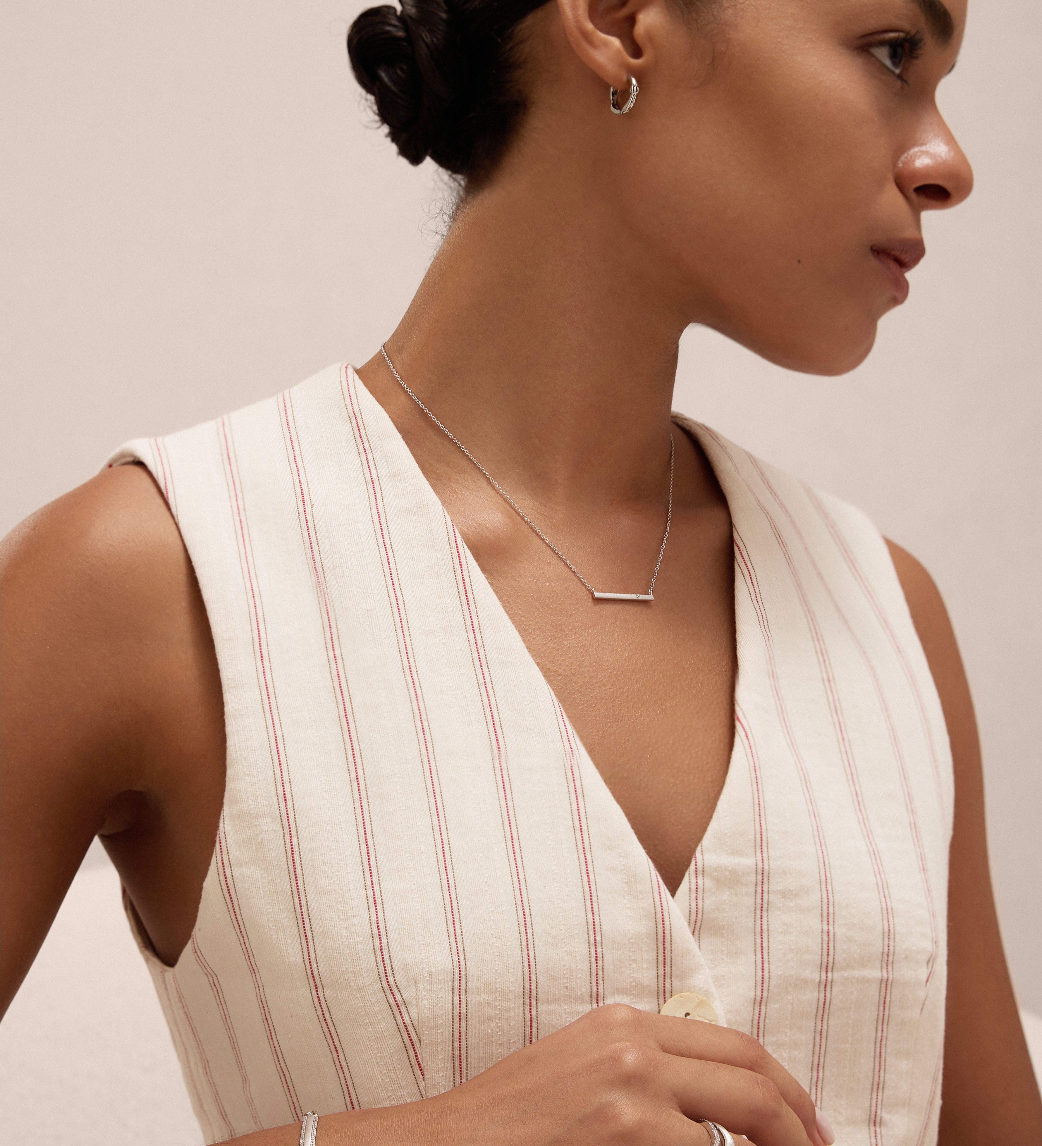 A woman with dark hair in a low bun models the Gisèle Cuban Link Bar Bracelet in Silver, paired with minimalist silver jewelry—hoop earrings, a bar necklace, and a simple ring—while wearing a cream pinstriped sleeveless top.