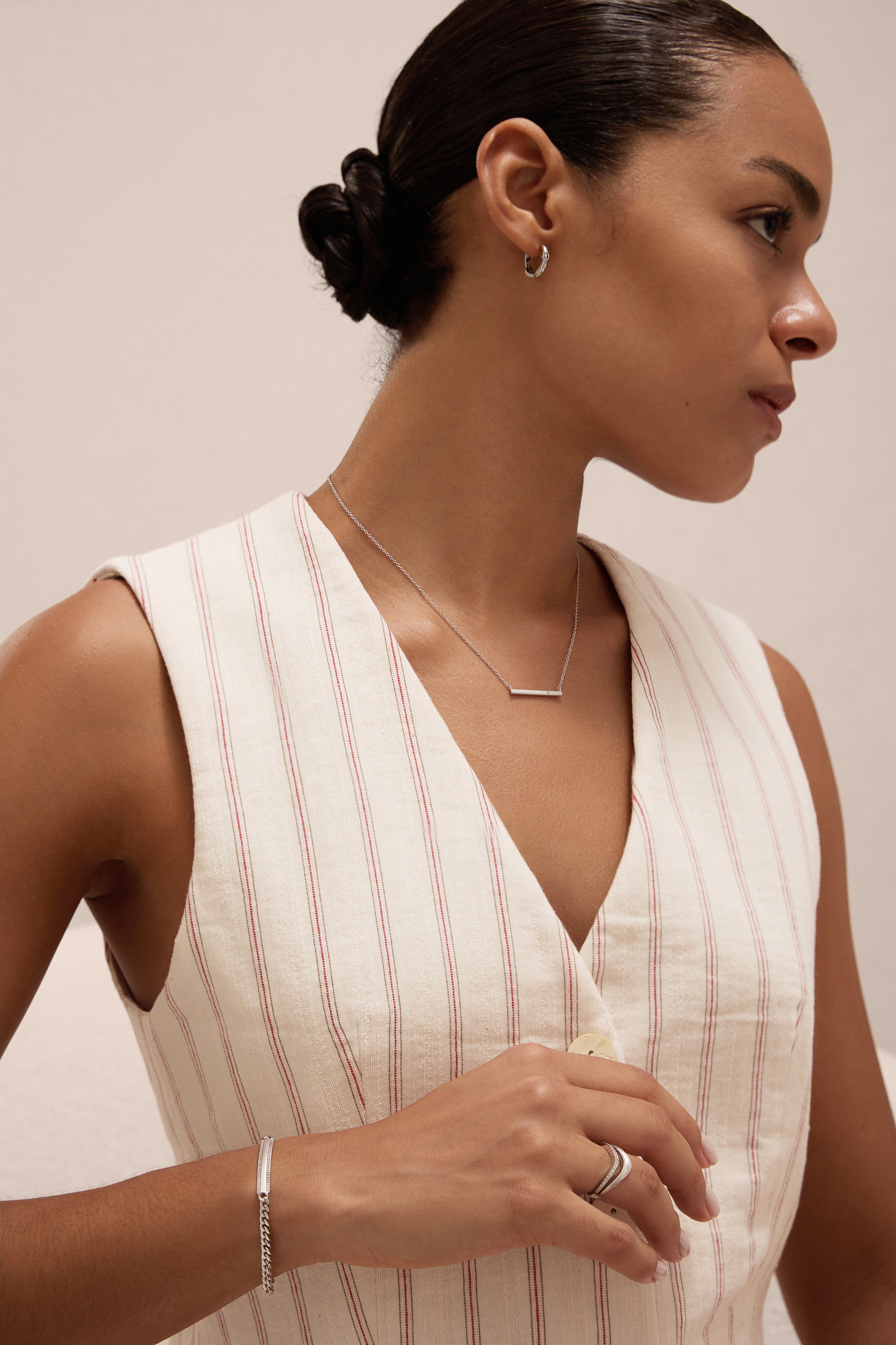 A woman with dark hair in a low bun models the Gisèle Cuban Link Bar Bracelet in Silver, paired with minimalist silver jewelry—hoop earrings, a bar necklace, and a simple ring—while wearing a cream pinstriped sleeveless top.