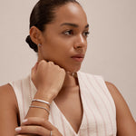 A woman with neatly tied-back hair wears a sleeveless, cream-striped top. Resting her chin on her hand, she showcases the sterling silver Gisèle Cuban Link Bar Bracelet along with rings and stud earrings against a neutral background.