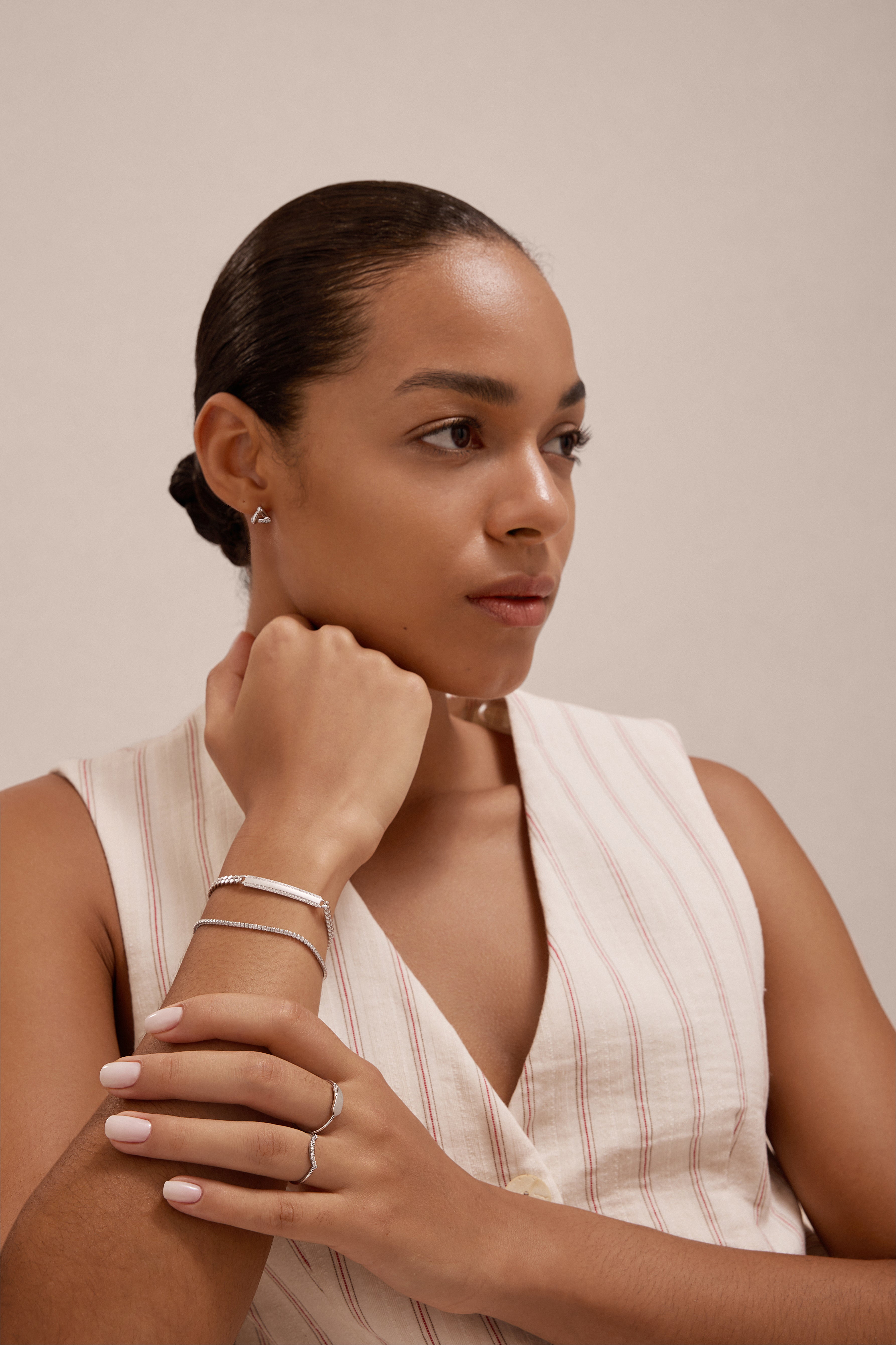 A woman with neatly tied-back hair wears a sleeveless, cream-striped top. Resting her chin on her hand, she showcases the sterling silver Gisèle Cuban Link Bar Bracelet along with rings and stud earrings against a neutral background.