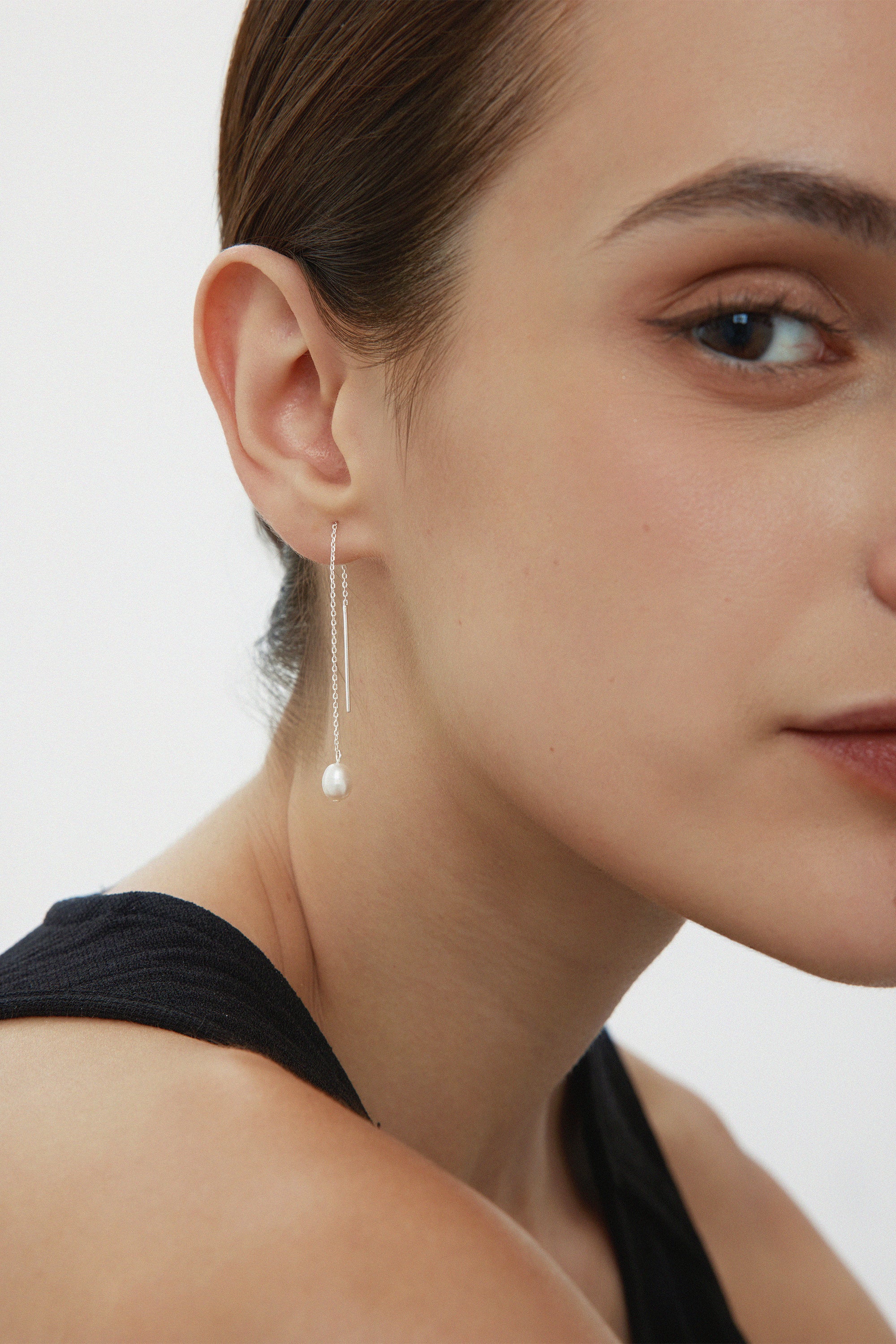 Close-up of a woman with brown hair in a black top, wearing Eira Pearl Drop Threader Earrings in Silver. She faces slightly right, highlighting her cheek, lips, and the elegant shimmer of her pearl drop earrings.