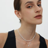 A woman with brown hair in a sleek updo wears the Estelle Herringbone Chain Necklace in Silver layered with another pendant and matching silver studs, paired with a black sleeveless top against a plain white background.