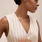 A woman with dark hair in a low bun wears a sleeveless cream top with red stripes and accessorizes with the Regina Horizontal Bar Pendant Necklace in Silver, embracing minimalist elegance against a light background.