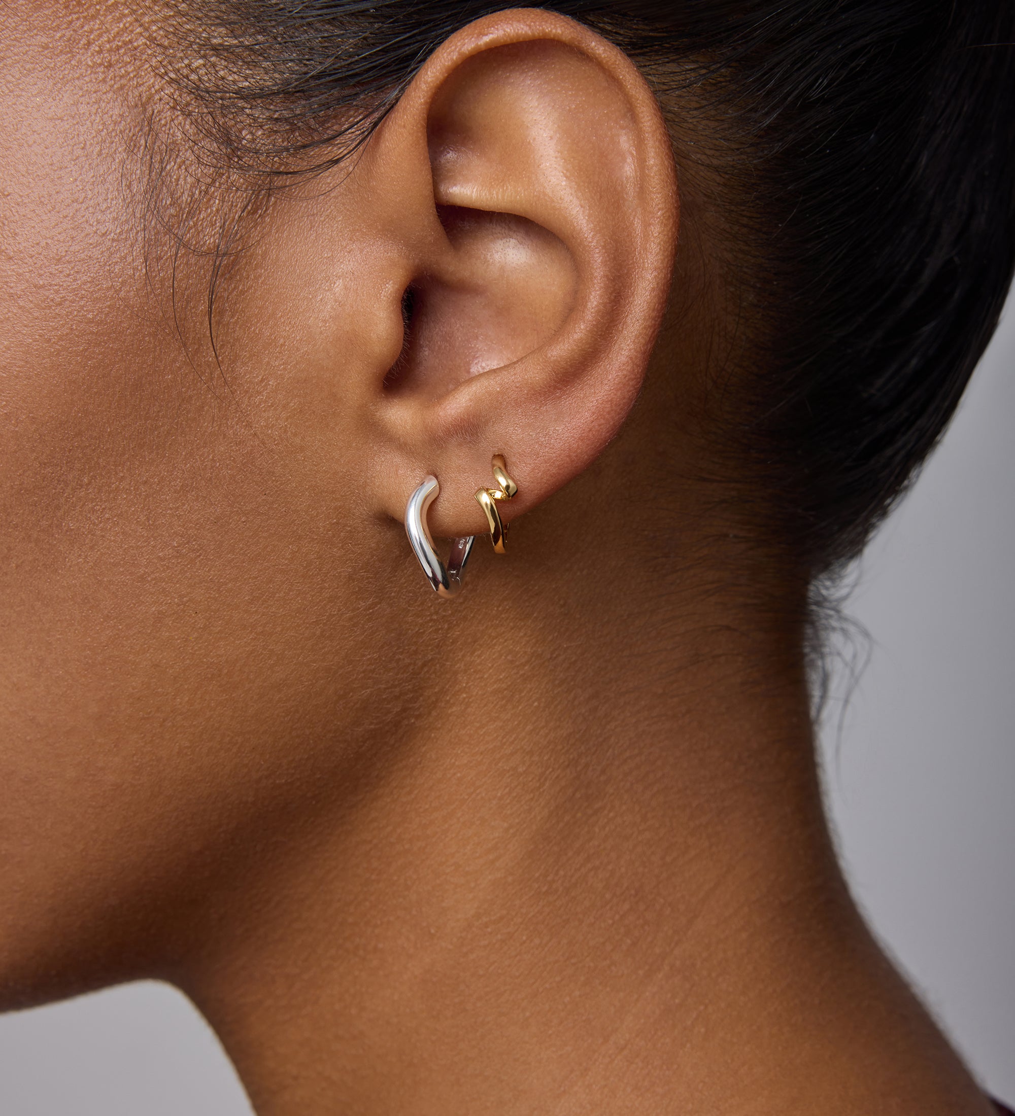 Close-up of a woman’s ear with everyday jewelry: a small gold hoop in the upper lobe and Nori Twisted Hoop Earrings in Silver in the lower lobe. She has smooth brown skin and dark hair pulled back.