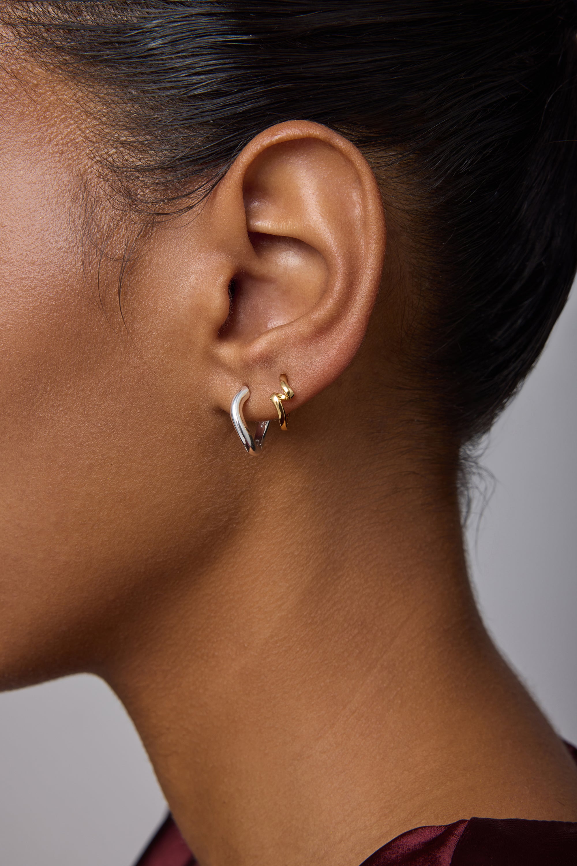 Close-up of a woman’s ear with everyday jewelry: a small gold hoop in the upper lobe and Nori Twisted Hoop Earrings in Silver in the lower lobe. She has smooth brown skin and dark hair pulled back.