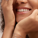 A smiling woman with curly hair rests her chin on one hand, partly covering her face with the other. She wears a sleeveless cream top, the Simone Triple Chain Layered Bracelet in Silver, simple silver rings, and other bracelets.