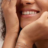 A smiling woman with curly hair rests her chin on one hand, partly covering her face with the other. She wears a sleeveless cream top, the Simone Triple Chain Layered Bracelet in Silver, simple silver rings, and other bracelets.