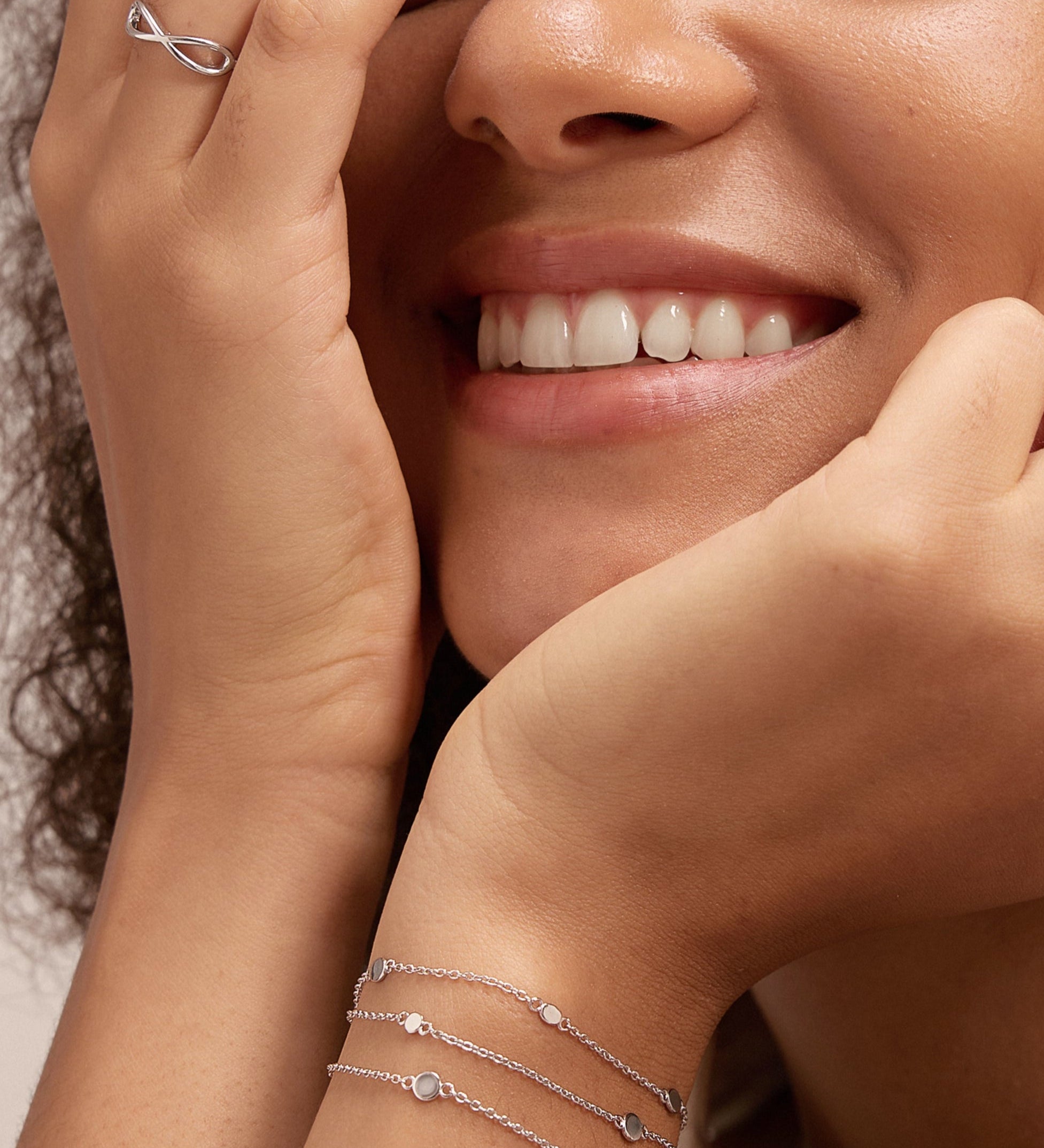 A smiling woman with curly hair rests her chin on one hand, partly covering her face with the other. She wears a sleeveless cream top, the Simone Triple Chain Layered Bracelet in Silver, simple silver rings, and other bracelets.