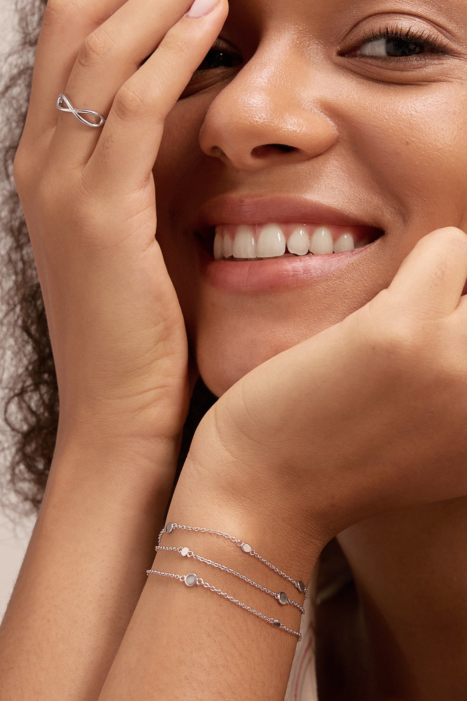 A smiling woman with curly hair rests her chin on one hand, partly covering her face with the other. She wears a sleeveless cream top, the Simone Triple Chain Layered Bracelet in Silver, simple silver rings, and other bracelets.