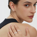 A woman with short, dark hair wears silver hoop earrings and the Vica Fluid Wave Chunky Band Ring in Silver, posing with her hand on her shoulder. She is dressed in a sleeveless black top against a plain white background.