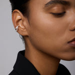 A close-up of a person with smooth, medium-brown skin and dark hair styled back, wearing a black blazer and the Vina Crystal Bezel Ear Cuff in Silver on their left ear. Only half of their face is visible.