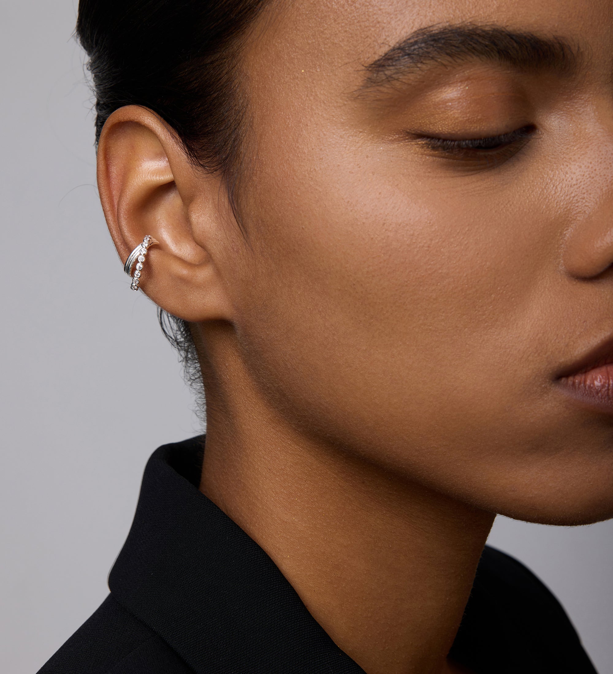 A close-up of a person with smooth, medium-brown skin and dark hair styled back, wearing a black blazer and the Vina Crystal Bezel Ear Cuff in Silver on their left ear. Only half of their face is visible.