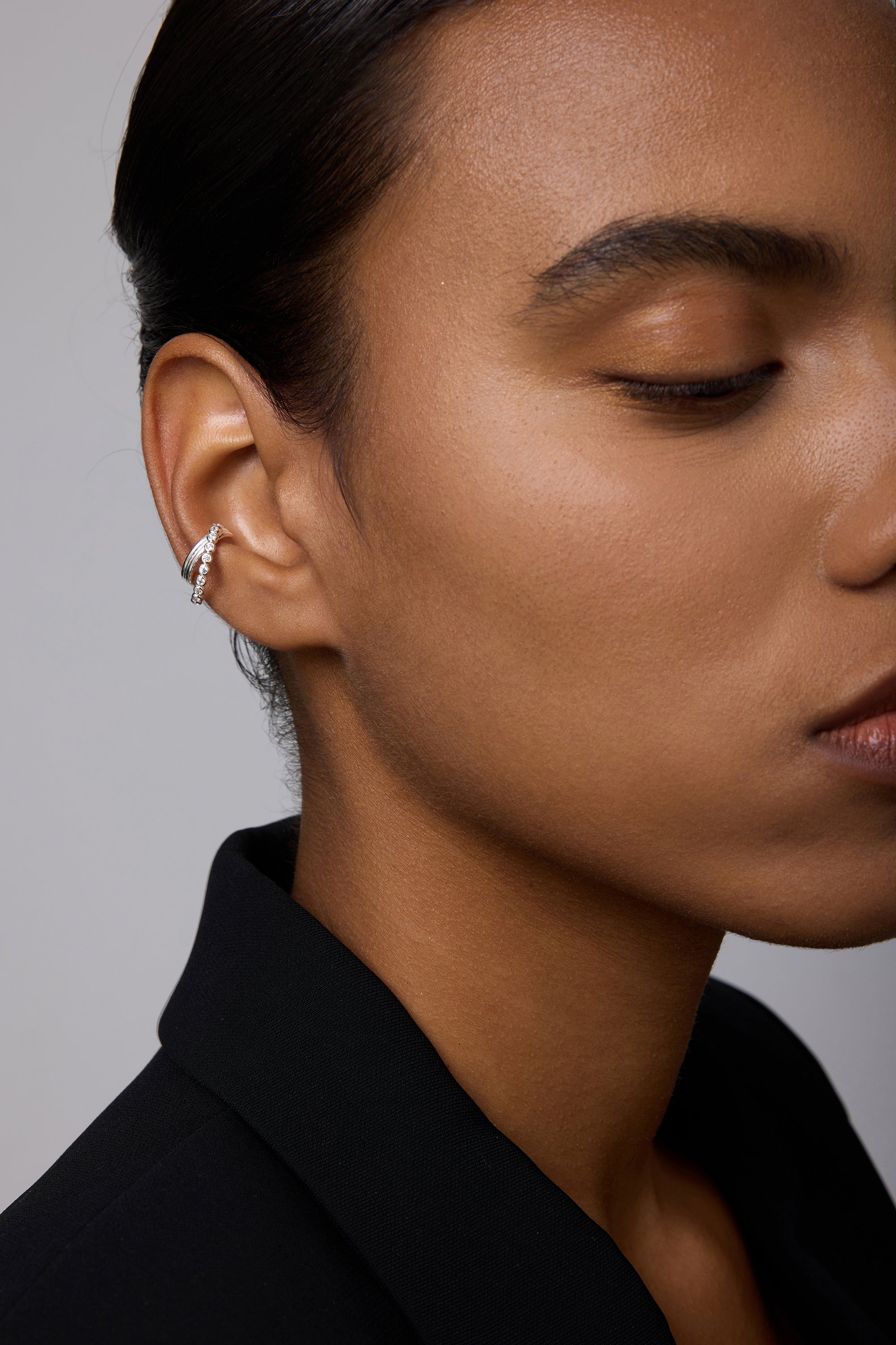 A close-up of a person with smooth, medium-brown skin and dark hair styled back, wearing a black blazer and the Vina Crystal Bezel Ear Cuff in Silver on their left ear. Only half of their face is visible.
