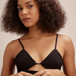 A woman with curly hair wears a black strappy top and delicate gold jewelry, including the Simone Triple Chain Layered Bracelet in Gold, standing against a neutral background and looking calmly at the camera with her hands gently clasped.