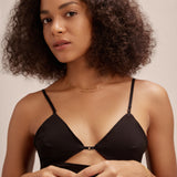 A woman with curly hair wears a black strappy top and delicate gold jewelry, including the Simone Triple Chain Layered Bracelet in Gold, standing against a neutral background and looking calmly at the camera with her hands gently clasped.