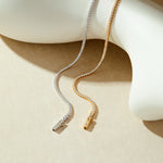 The Alba Tennis Necklace in Gold, adorned with sparkling cubic zirconia, drapes elegantly over a smooth, white abstract sculpture on a beige background.
