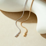 The Alba Tennis Necklace in Gold, adorned with sparkling cubic zirconia, drapes elegantly over a smooth, white abstract sculpture on a beige background.