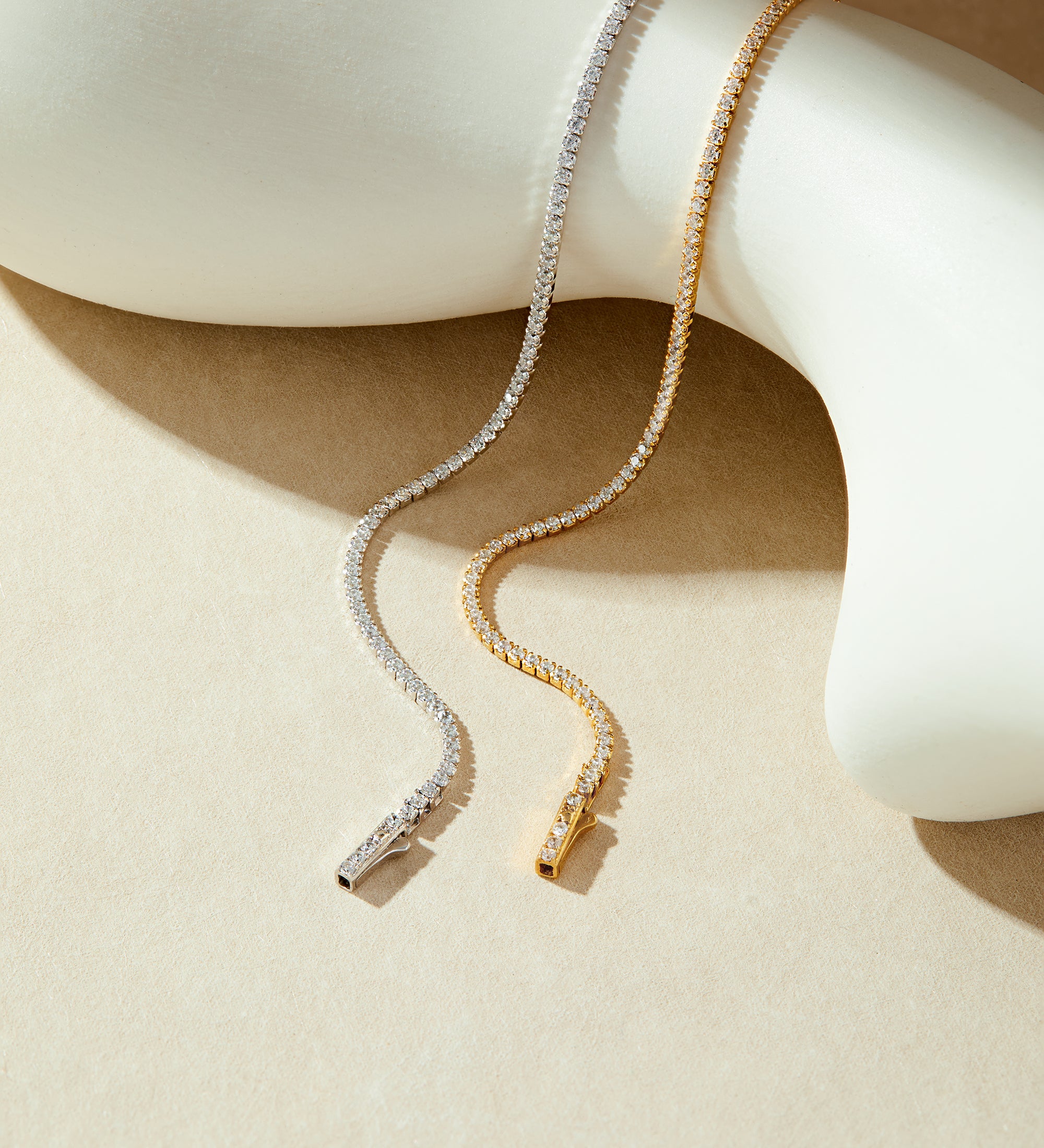 The Alba Tennis Necklace in Gold, adorned with sparkling cubic zirconia, drapes elegantly over a smooth, white abstract sculpture on a beige background.