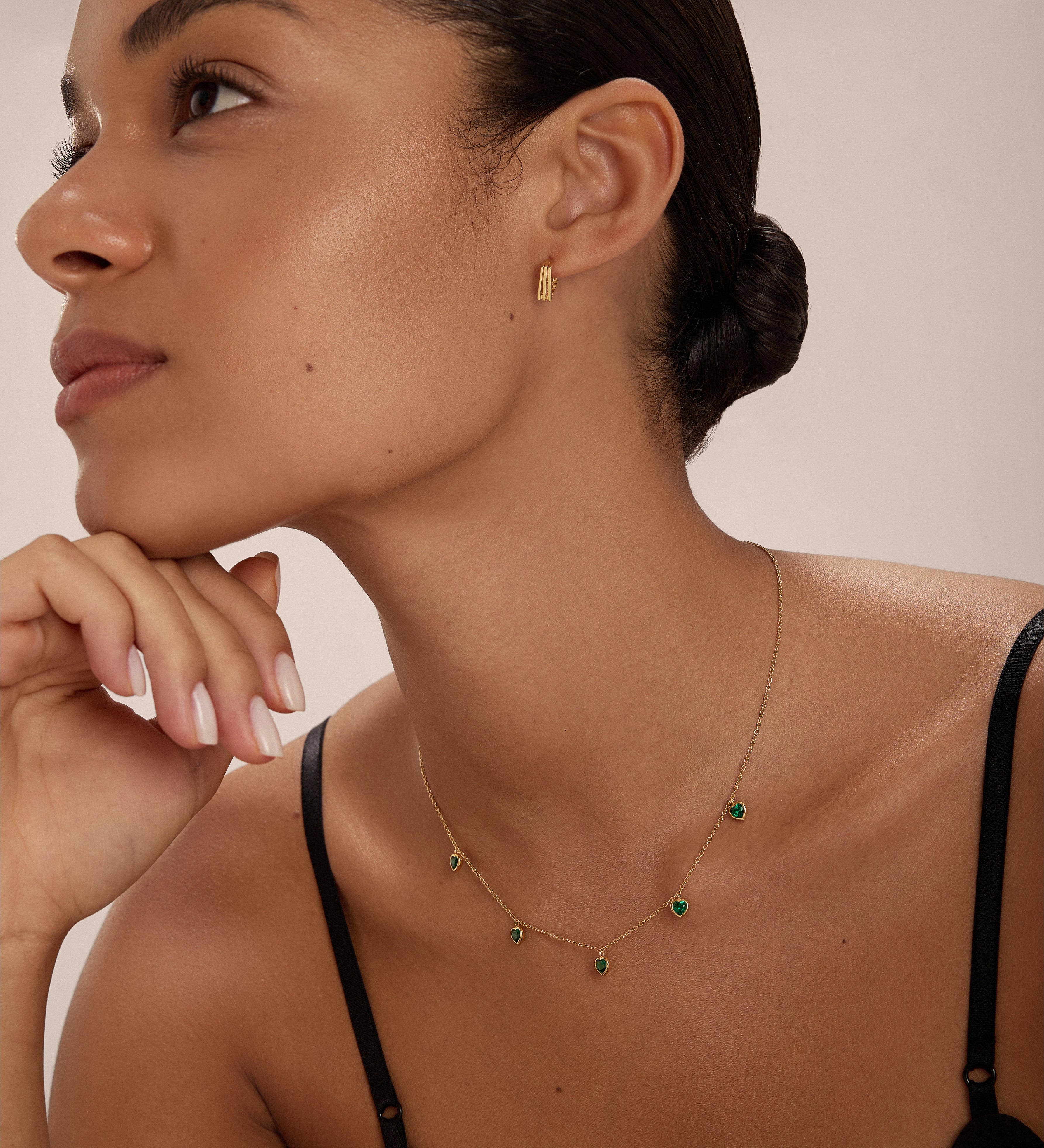 A woman with smooth, glowing skin and dark hair tied back wears the Thea Green Heart Pendant Gold Necklace with matching gold earrings. She poses in a black spaghetti strap top against a neutral background.