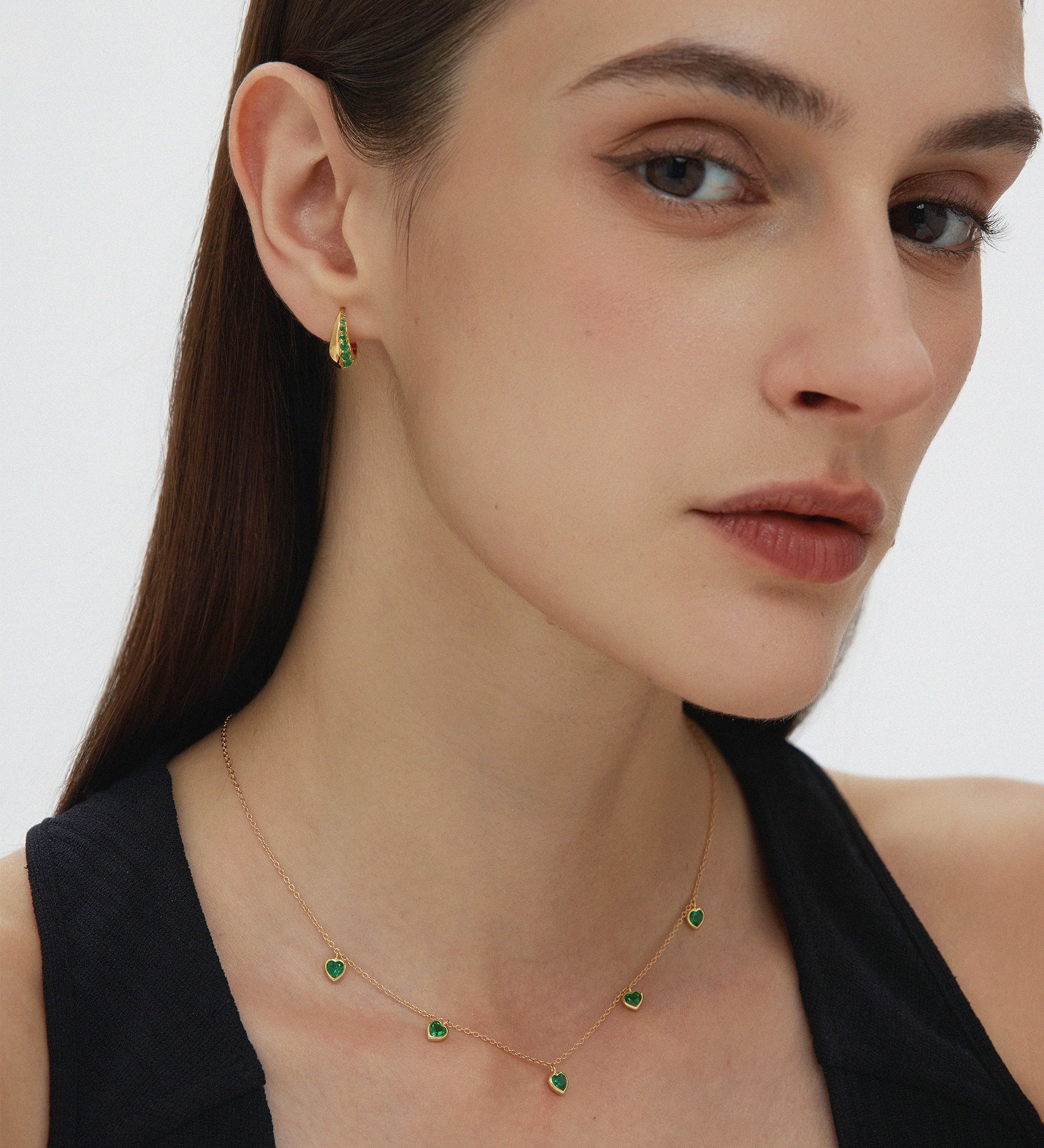 A woman with straight brown hair wears a sleeveless black top, small green gemstone earrings, and the Thea Green Heart Pendant Gold Necklace. She looks slightly to the side with a neutral expression.